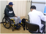 車椅子使用者の横に介助犬が座り、医師による定期受診を受けている様子