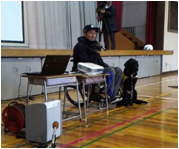 小学校の体育館のステージ前で、車いすユーザーが講演をしている間、介助犬が横でおとなしく座っている様子